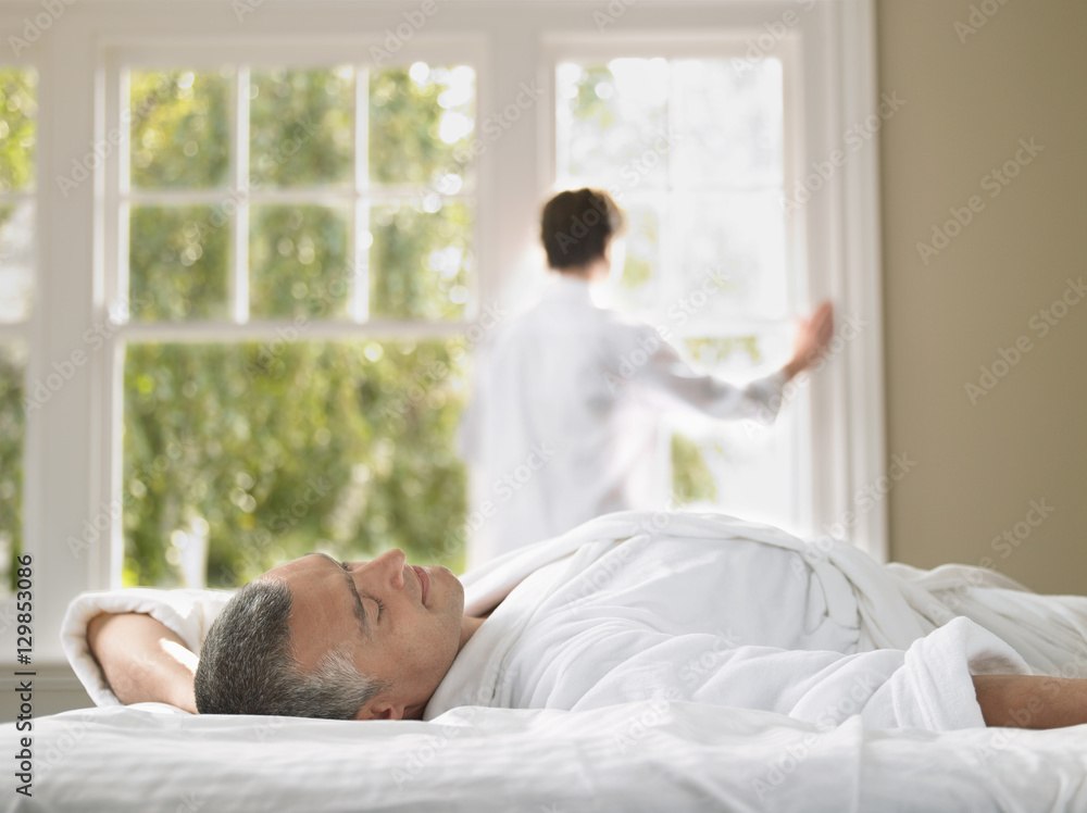 Man wearing bathrobe lying on bed with woman standing against window in bedroom Man wearing bathrobe lying on bed with woman standing against window in bedroom