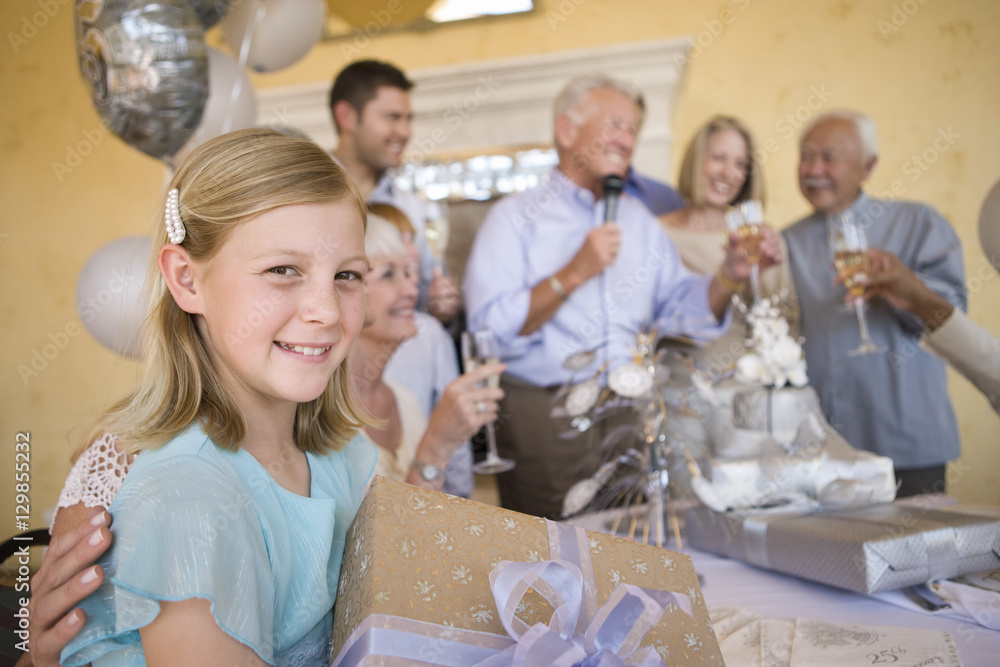 Portrait of teenage girl holding gift with family celebrating in the background Portrait of teenage girl holding gift with family celebrating in the background
