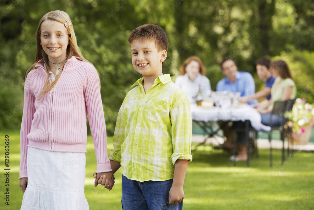 Portrait of happy brother and sister holding hands with family having lunch in background Portrait of happy brother and sister holding hands with family having lunch in background