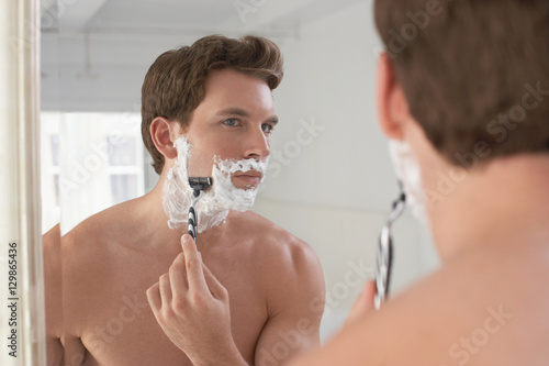 Reflection of a young man shaving in bathroom mirror
