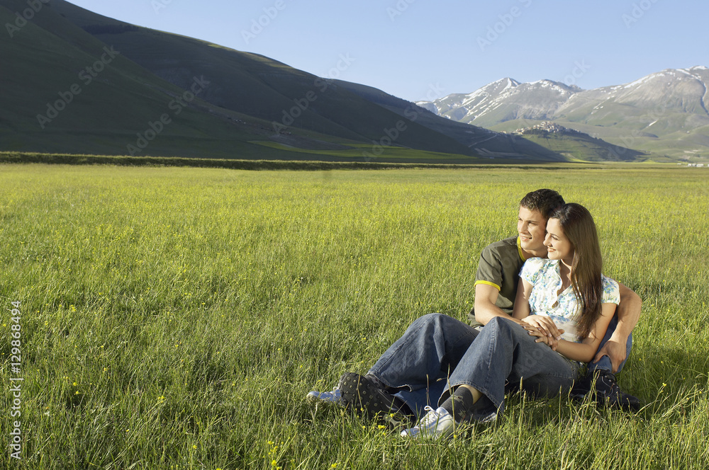 Young loving couple looking away while sitting together on grassy field at against mountain at park