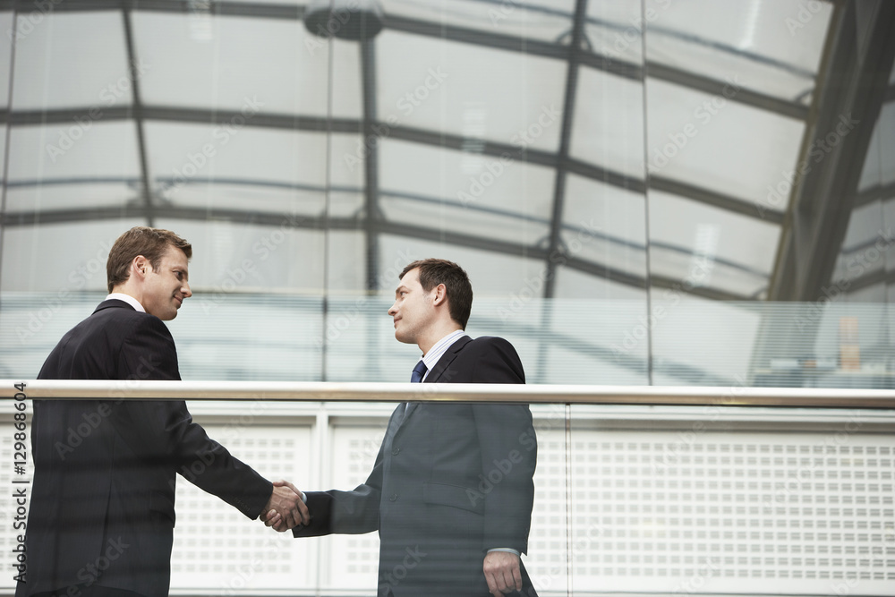 Businessmen shaking hands while standing against glass railing in office Businessmen shaking hands while standing against glass railing in office