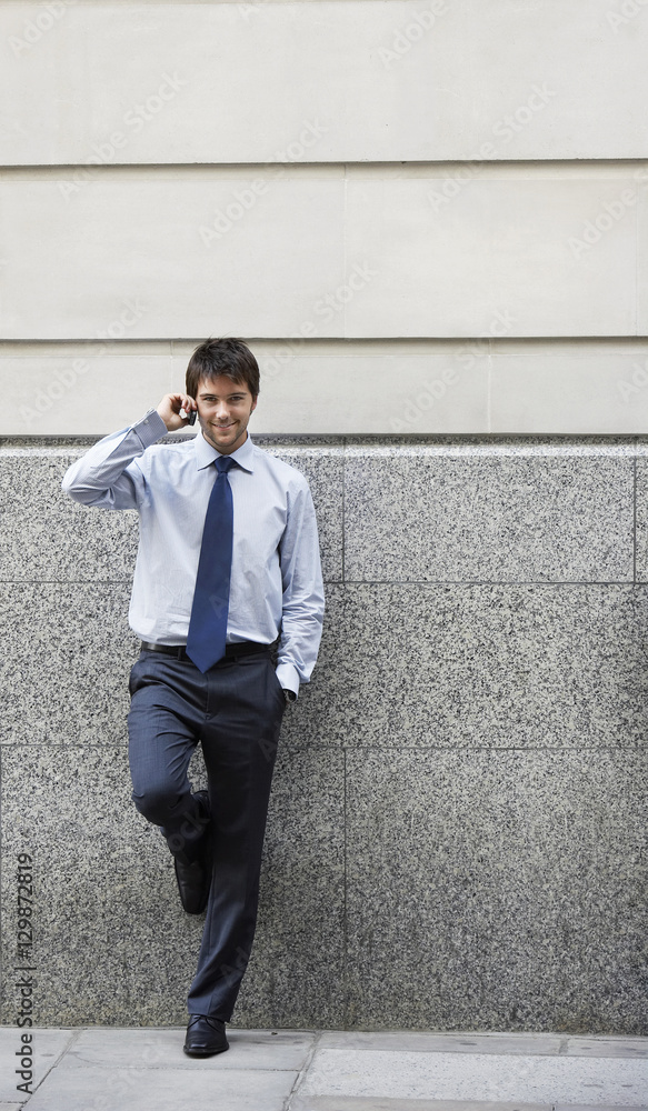 Full length portrait of happy young businessman on call leaning against wall Full length portrait of happy young businessman on call leaning against wall