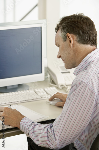 Middle aged male businessman using computer while noting on paper at desk