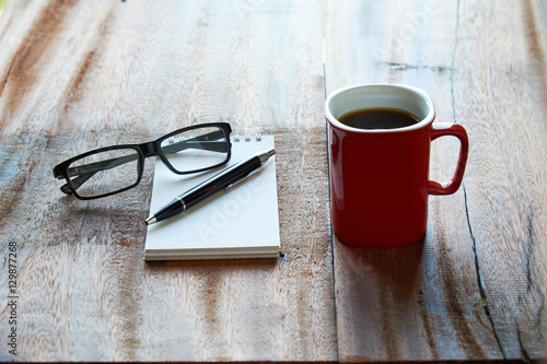 A cup of coffee with notepad, pen and glasses on wooden table. idea of writing and thinking while enjoy a coffee.