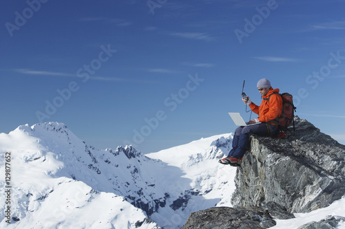 Mountain climber using laptop with walkie talkie on mountain peak against blue sky photo