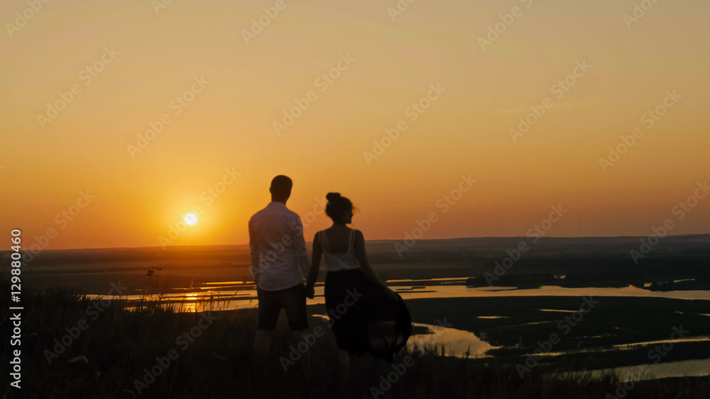 loving couple - brave young man and beautiful girl stands on high hill  looking to sunset silhouette