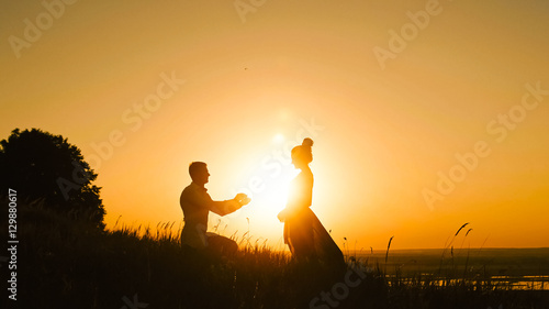 Romantic Silhouette of Man Getting Down on his Knee and Proposing to Woman high hill - Couple Gets Engaged at Sunset - Putting Ring Girl s Finger