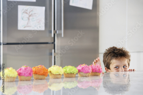 Happy young boy peaking over counter at row of cupcakes in kitchen