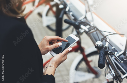 Young businesswomen in black suit and umbrella using smartphone, biking and going to work by city bicycle on urban street, hipster girl holding mobile gadget, ecology environment concept