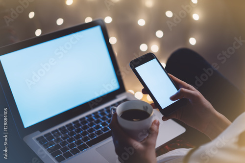 Hipster girl using computer and cup coffee in home atmosphere, person holding smartphone on background glow bokeh Christmas illumination, female hands texting on glitter decoration, mockup, blur