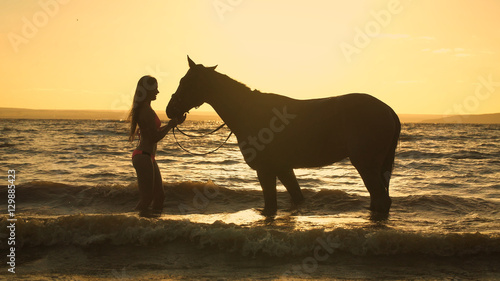 A beautiful blonde young sexy woman standing next to a horse at a lake