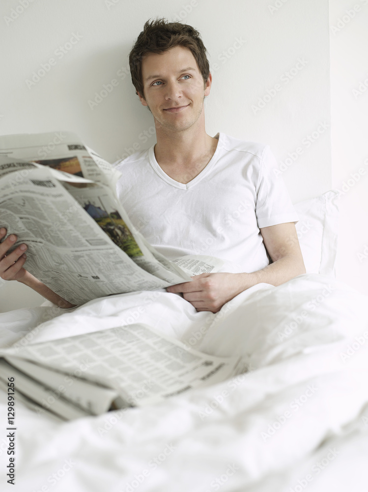 Happy young man with newspaper in bed looking away Happy young man with newspaper in bed looking away