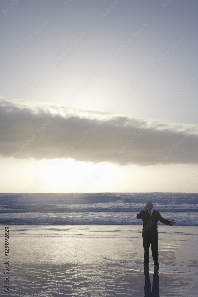 Middle aged businessman using cell phone in sea at sunset