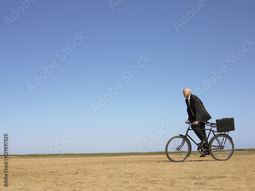 Side view of middle aged businessman with briefcase riding bicycle in desert © moodboard