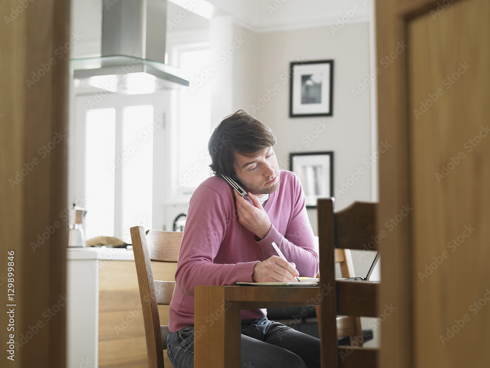Young man writing notes while communicating on mobile phone at kitchen table Young man writing notes while communicating on mobile phone at kitchen table