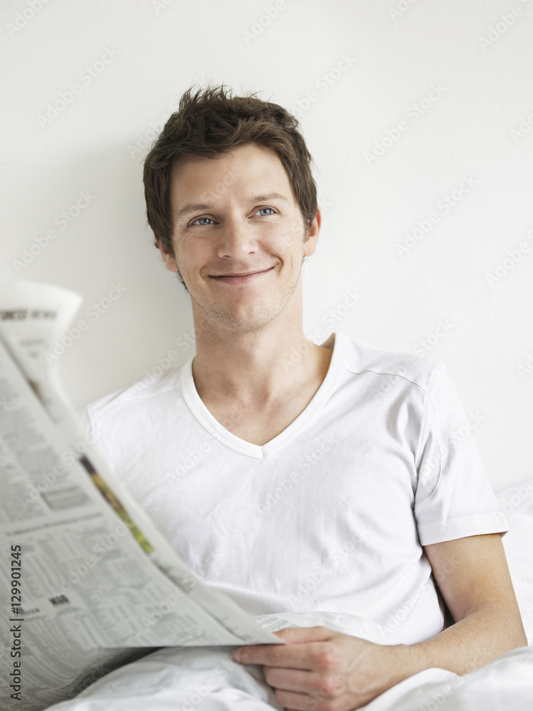Smiling young man with newspaper in bed looking away Smiling young man with newspaper in bed looking away