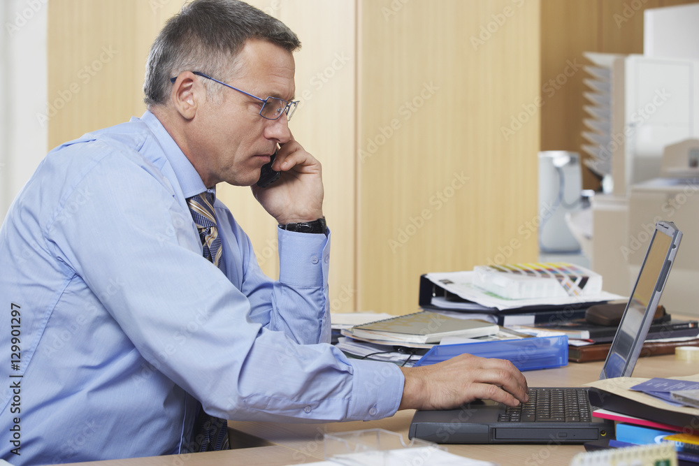 Side view of middle aged businessman on call while using laptop at desk