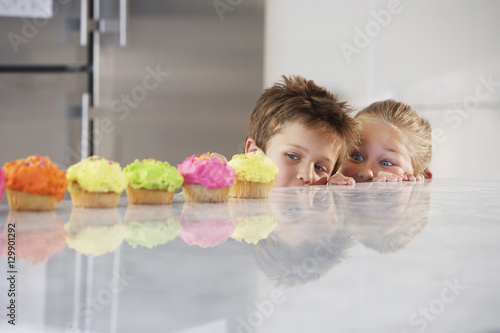 Young girl and boy peeking over counter at row of cupcakes