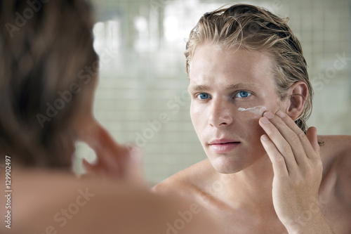 Closeup of handsome young man applying facial cream in front of mirror photo