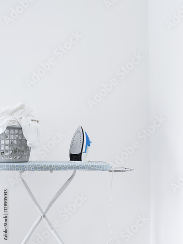 Iron and laundry basket on ironing board against white wall photo