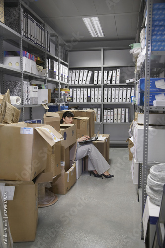 Foto Side view of a young woman using laptop between boxes in storage room