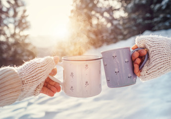 Man and woman hands in knitting mittens with cups of hot tea on winter forest glade