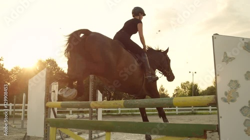 CLOSE UP: Young woman rider and a horse jumping over a fence on arena at sunset