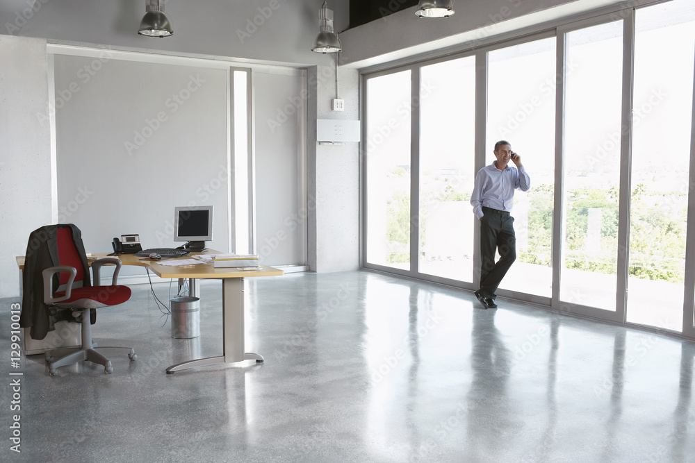 Full length of a male executive using cellphone against glass wall in empty office Full length of a male executive using cellphone against glass wall in empty office