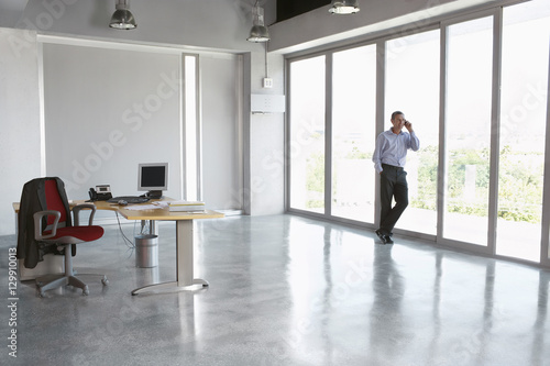 Full length of a male executive using cellphone against glass wall in empty office