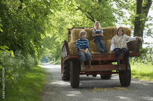 Full length portrait of three young kids sitting on back of trailer on country lane