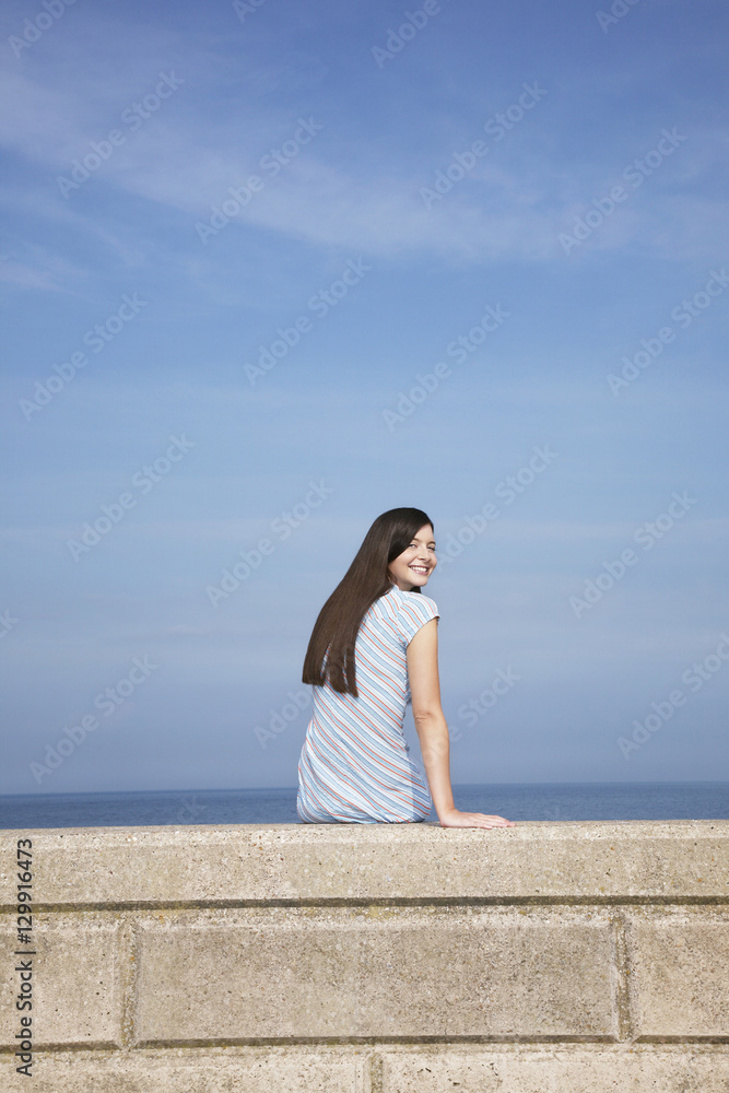Rear view portrait of happy young woman sitting on stone ledge at beach