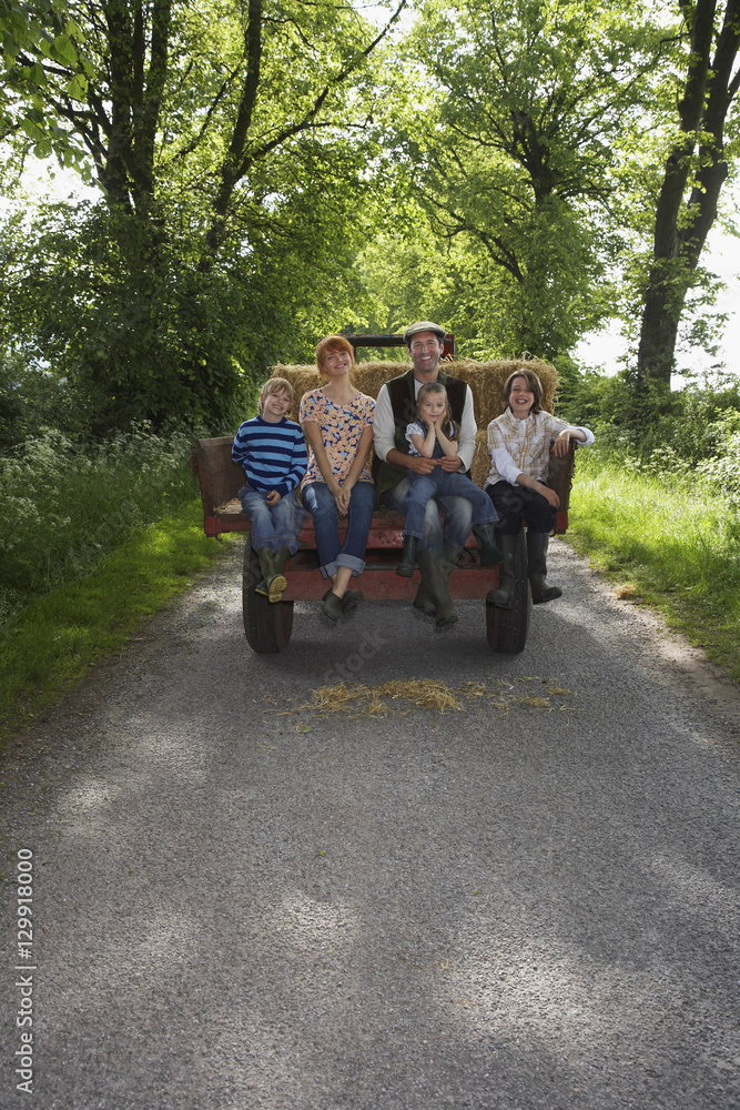 Full length portrait of couple with three kids sitting on back of trailer on country lane Full length portrait of couple with three kids sitting on back of trailer on country lane