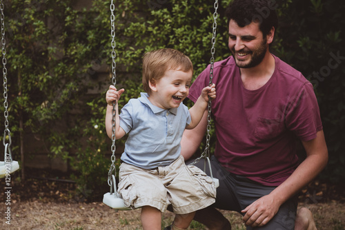 Smiling father twisting swing with little boy and laughing on background of green fence. © progat