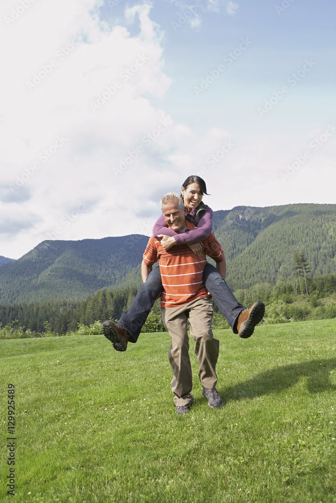 Full length of an active middle aged man carrying woman on back on meadow
