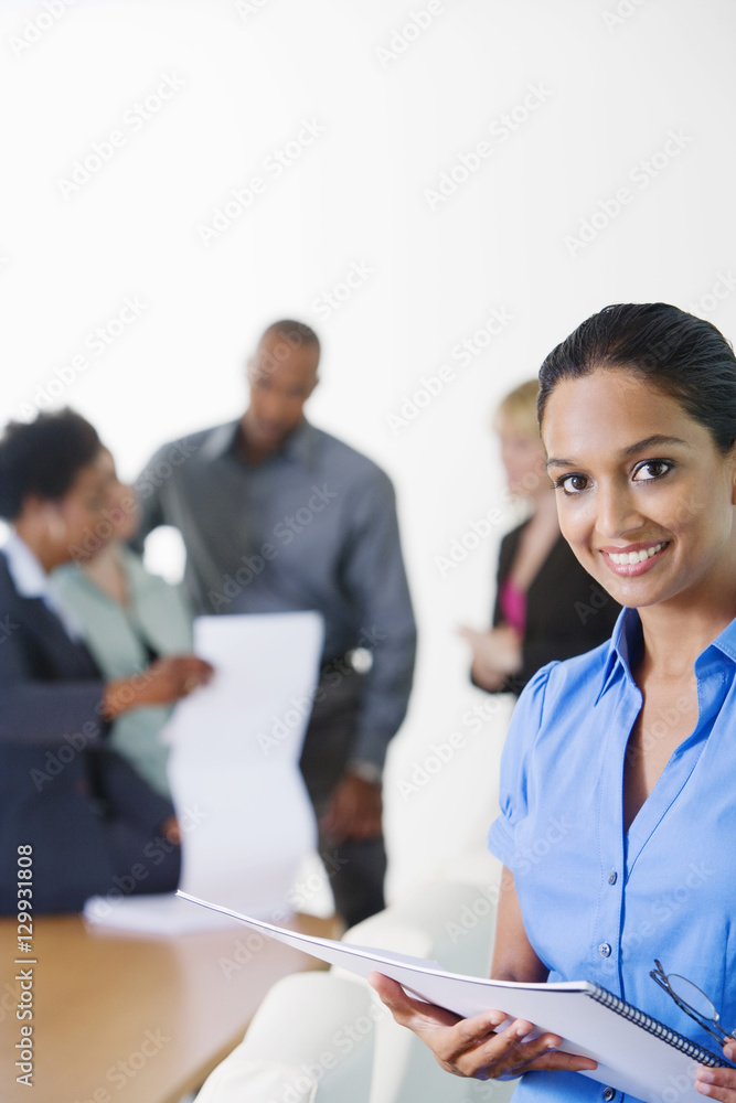 Portrait of a female estate agent holding notepad with colleagues discussing in the background Portrait of a female estate agent holding notepad with colleagues discussing in the background
