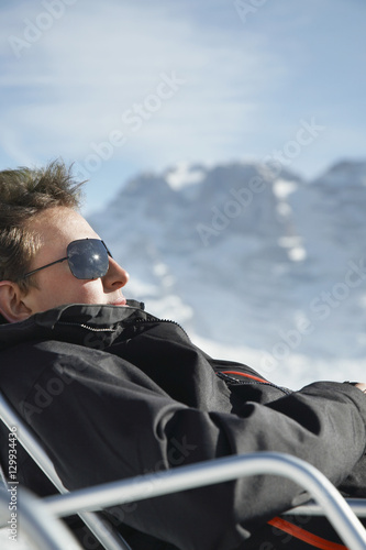 Side view of young man in winter clothes relaxing on chair against mountain