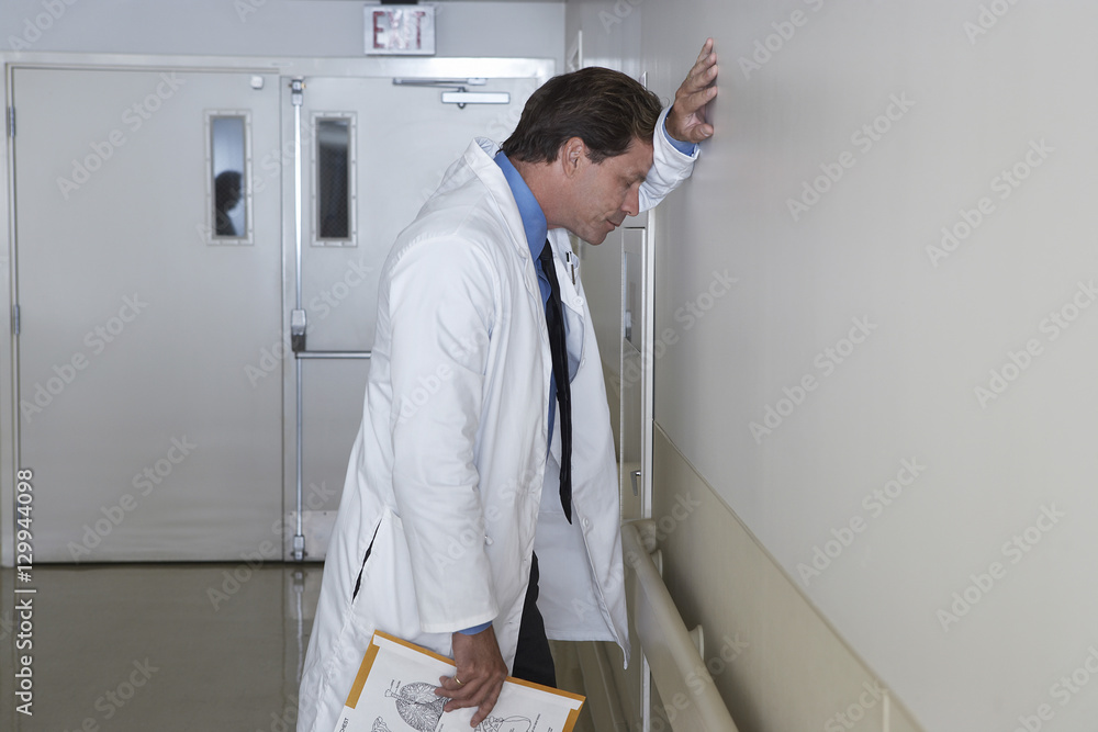 Side view of a depressed doctor leaning against wall in hospital corridor