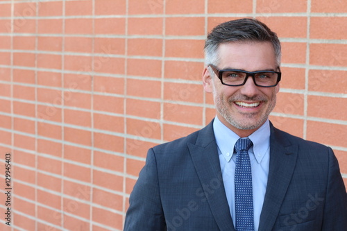 Confident and successful. Cheerful mature man in suit, shirt and tie looking at camera and keeping arms crossed while standing against brick wall urban background