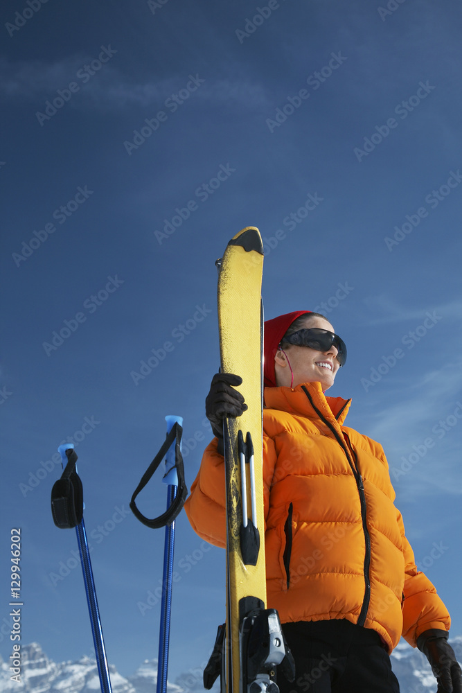 Naklejka premium Low angle view of happy woman in winter jacket holding ski against blue sky