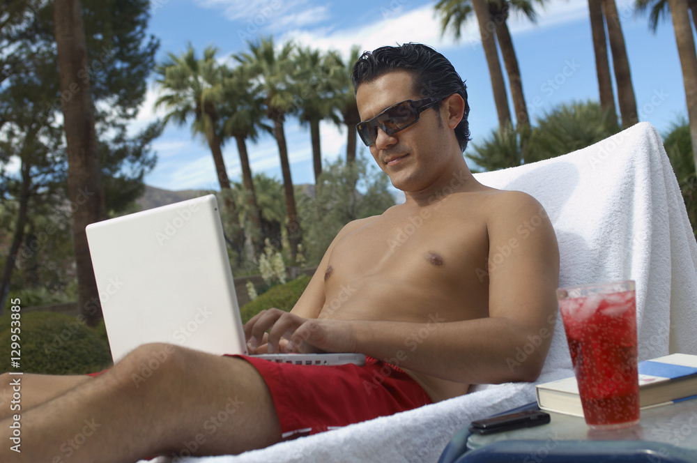 Shirtless man using laptop while sitting on lounge chair outdoors