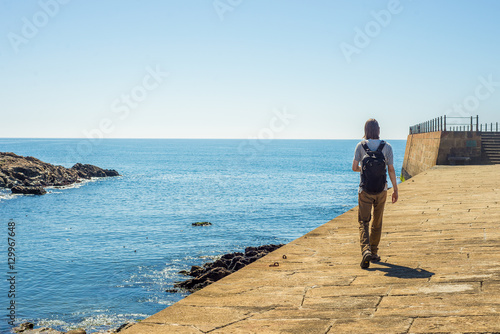 Tourist man walking harbor pier seafront near ocean on sunny day wearing black backpack