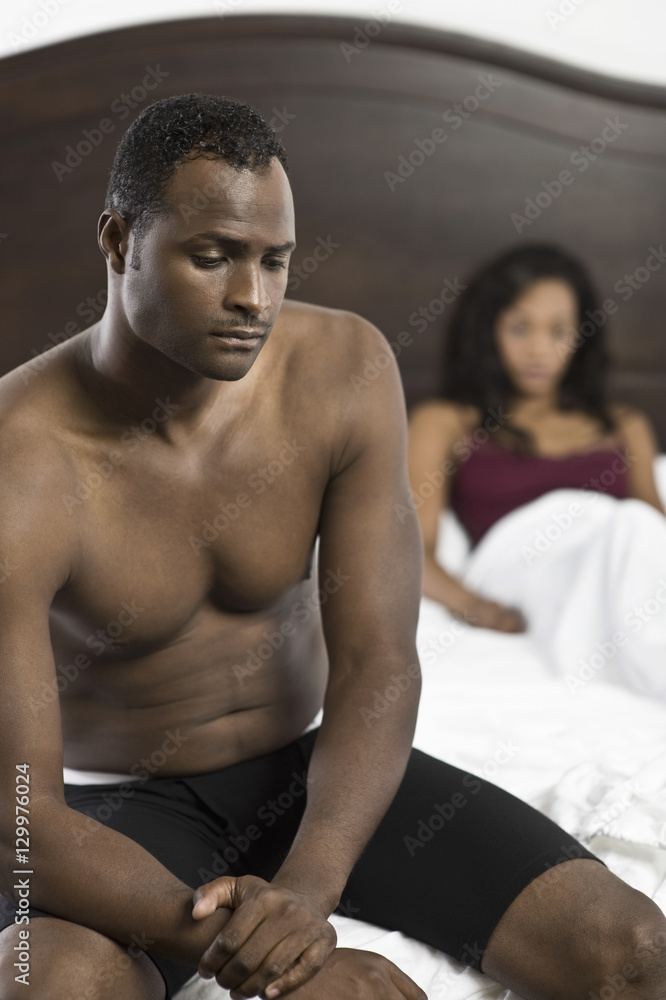 Tensed shirtless African American man sitting on bed with blurred woman in background Tensed shirtless African American man sitting on bed with blurred woman in background