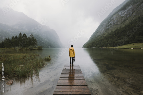 Young adult male wearing yellow jacket standing on a footbridge at a foggy mountain lake with forest 