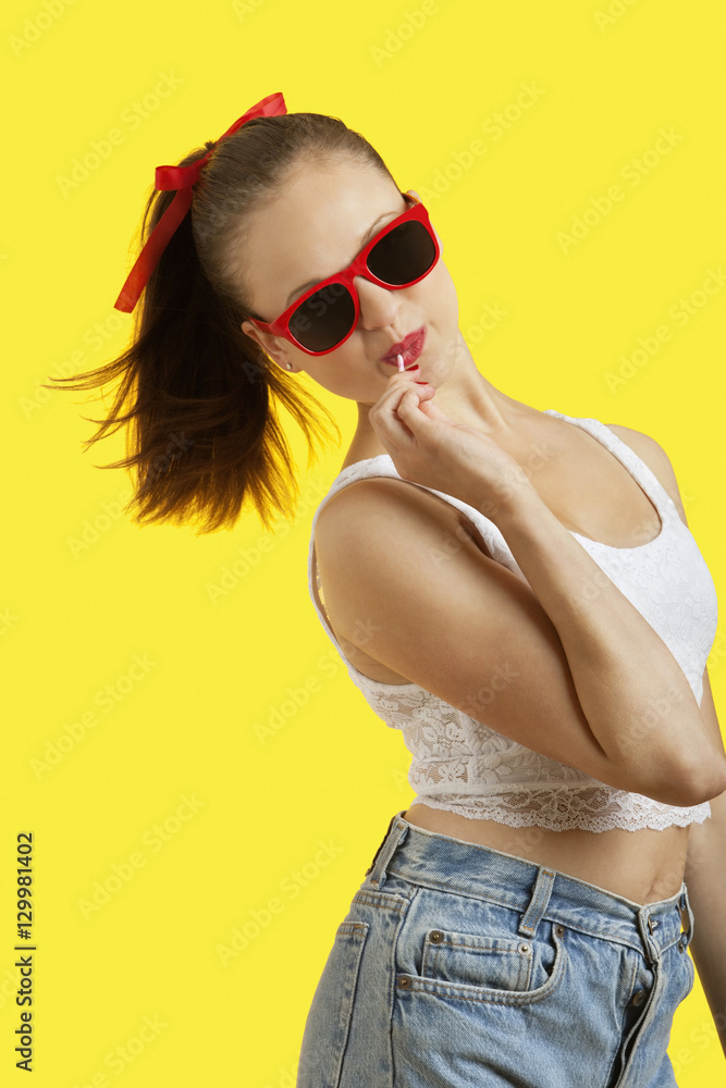 Portrait of playful young woman in sunglasses eating lollipop over yellow background