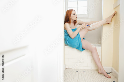 Young female shaving her legs on the side of the bathtub