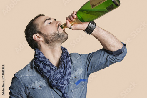 Young man drinking champagne from bottle over colored background