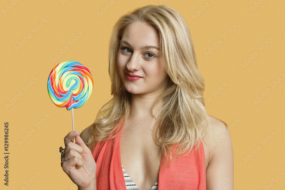 Portrait of a young blond woman holding lollipop over colored background