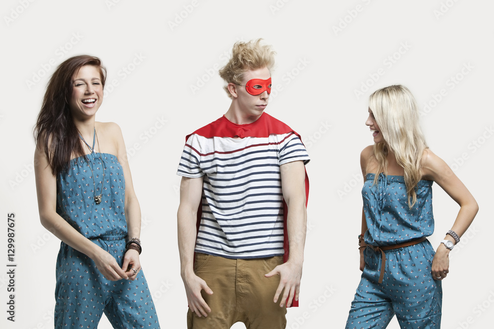 Young man in superhero costume standing with happy female friends in matching jump suits against gray background Young man in superhero costume standing with happy female friends in matching jump suits against gray background