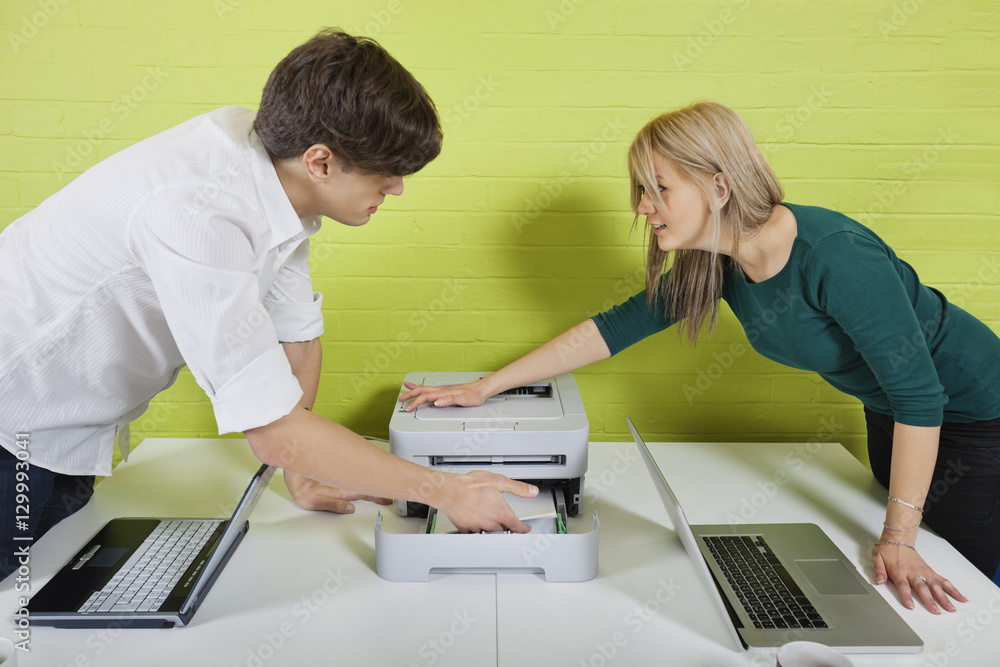 Side view of young businesspeople setting up printer with laptops at desk Side view of young businesspeople setting up printer with laptops at desk